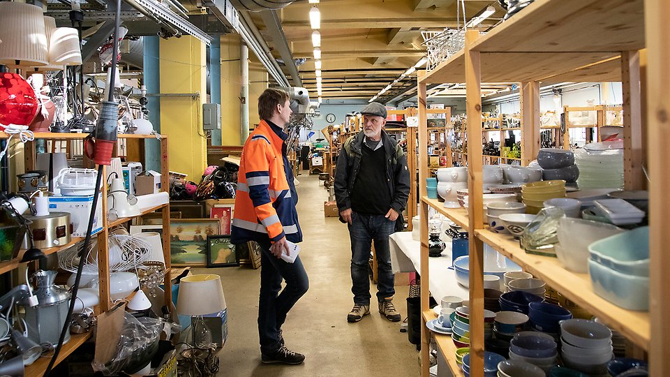 Bild: Två män står bland hyllor fyllda med prylar och pratar