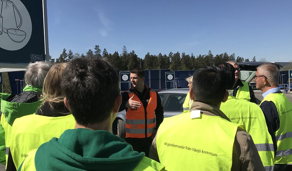 Politiker i gula västar står framför personal från SSAM på återvinningscentralen.